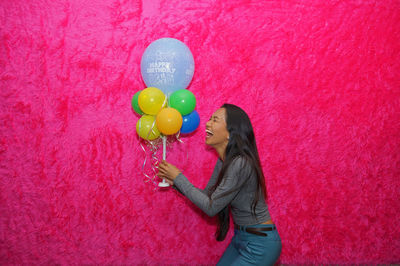 Side view of cheerful young woman holding birthday balloons against pink velvet