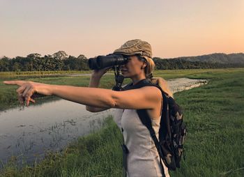 Woman standing on field while looking through binoculars against sky during sunset