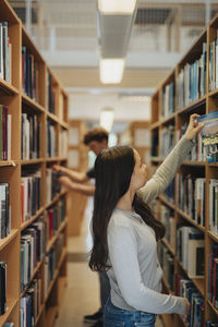 Side view of female student searching books on bookshelf in library at university