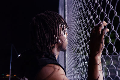 Rear view of young man looking through chainlink fence