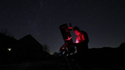 Man using mobile phone by telescope against sky at night