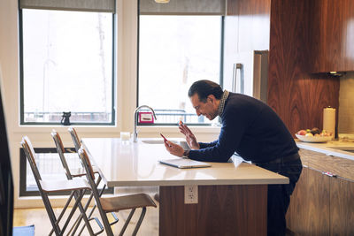 Man waving while using smart phone at kitchen table in home