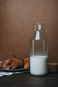 Glass decanter with milk and plate with fresh croissants, brown background. 