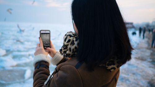 Rear view of woman photographing through mobile phone during winter at beach