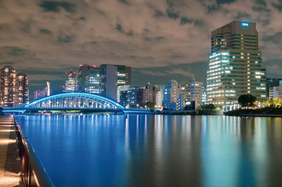 Illuminated modern buildings by river against sky at night