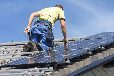 Low angle view of man installing solar panels on roof against sky