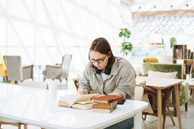 Young teenager girl college student in glasses doing homework with books at green modern library 