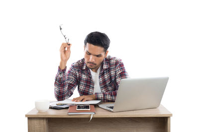 Businessman holding eyeglasses while reading document at laptop desk against white background