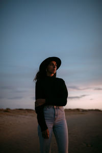 Pensive young female in casual clothing and hat thoughtfully looking away in empty meadow under light blue pink sky with clouds at dawn