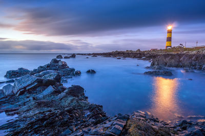 Illuminated buildings by sea against sky during sunset