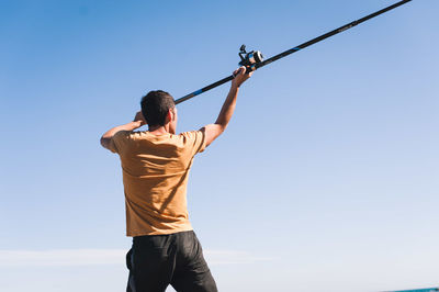 Rear view of man holding fishing rod while standing against sky during sunny day