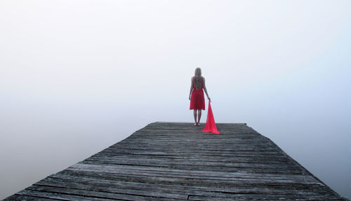 Rear view of woman holding scarf while standing on wooden pier by lake in foggy weather