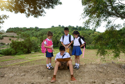 Siblings standing by brother reading while sitting on field against trees