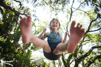 Low angle portrait of cheerful girl swinging in forest