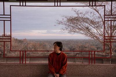 Young man sitting on seat against cloudy sky during sunset