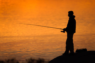 Silhouette man fishing in sea against orange sky