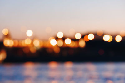 Defocused image of illuminated lights by sea against sky at sunset