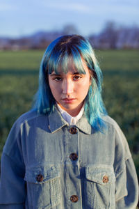 Thoughtful young female with blue hair looking at camera dressed in trendy jacket standing in green field in sunny evening