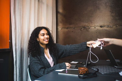 Smiling young female professional giving charger to male colleague at desk in office