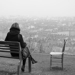 Full length rear view of woman sitting on bench by cityscape during foggy weather