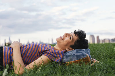 Side view of happy woman lying on grassy field against cloudy sky in city