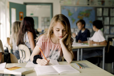 Female student with hand on chin studying from book while sitting in classroom