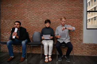 Business people waiting while sitting on chairs against brick wall in office