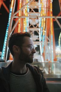 Young man looking away while standing against illuminated ferris wheel at night