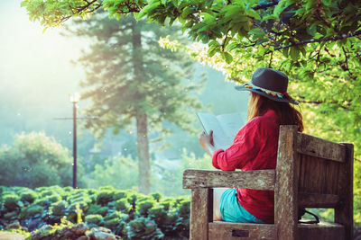 Woman wearing hat reading book while sitting on bench at park