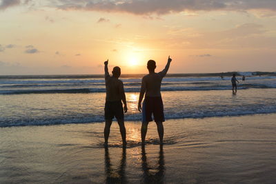 Rear view of shirtless men pointing at sea while standing on shore at beach against sky during sunset