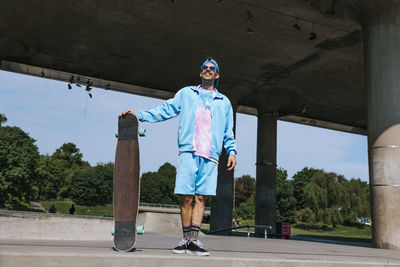 Smiling fashionable man in casuals standing with longboard near overpass at sunny day