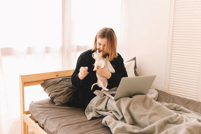 A cheerful young teenage woman plays with her pet a small dog and works using a laptop at home