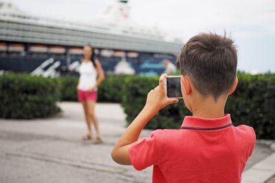 Rear view of son photographing mother with smart phone at park