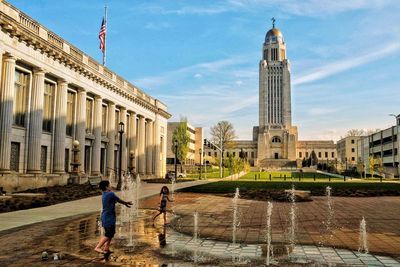 Cheerful boy and girl standing at fountain in city against sky