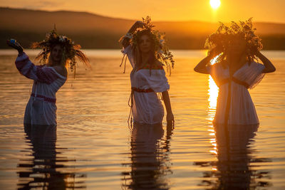 Rear view of women standing at sea shore against sunset sky