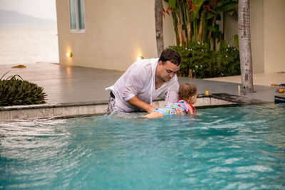 Father teaches cute toddler little girl in swimsuit to swim in the pool holding his hand.