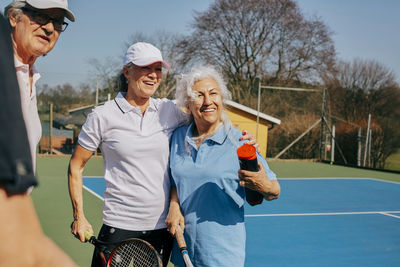 Happy senior friends looking away while holding tennis racket at court
