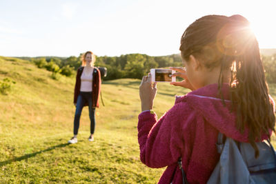 Rear view of girl photographing mother standing on field against clear sky during sunset
