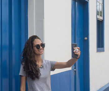 Smiling young woman taking selfie while standing by wall