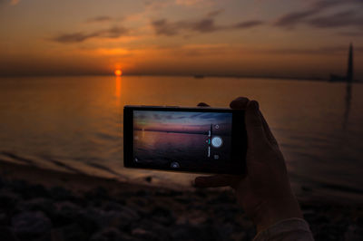 Cropped hand of person photographing sea with smart phone against sky during sunset