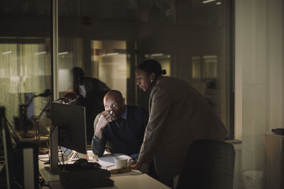 Mature businessman drinking coffee while discussing with female colleague over computer in office