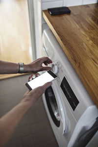 Cropped hands of senior woman holding mobile phone while using washing machine in bathroom at home