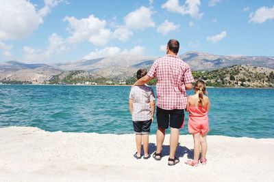 Rear view of father with children standing on shore at beach against sky