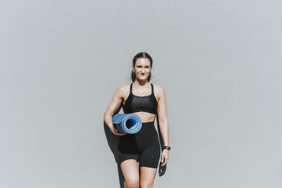 Smiling beautiful young woman with exercise mat leaning on wall