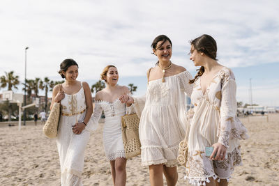 Young friends in white dresses smiling while walking at beach