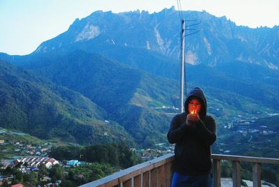 Man with hooded shirt igniting cigarette by railing against mountains