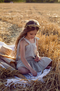 Girl child in a dress and a wreath on her head sit on a mown field of wheat at sunset in summer