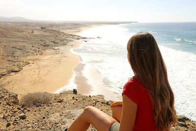 Rear view of woman sitting on cliff against sea and sky