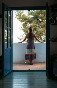 Full body back view of unrecognizable female in casual sundress admiring view from balcony of country house while spending summer holidays in tropical countryside