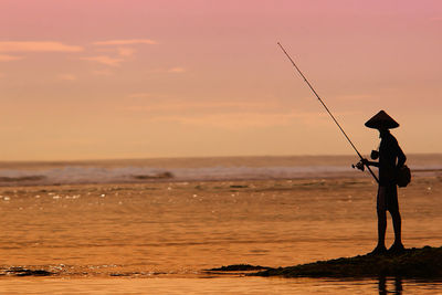 Man fishing on beach against sky during sunset